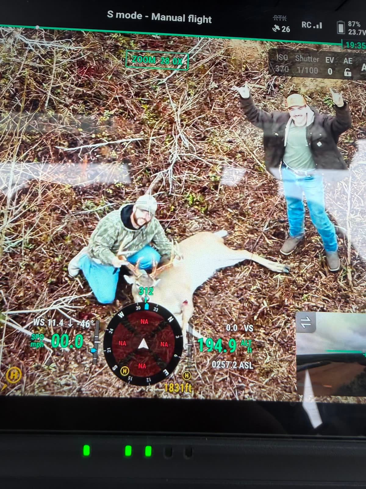 Drone camera looking down on two hunters in a cutover beside the recovered buck, one with both arms raised, flight telemetry overlaid on the frame